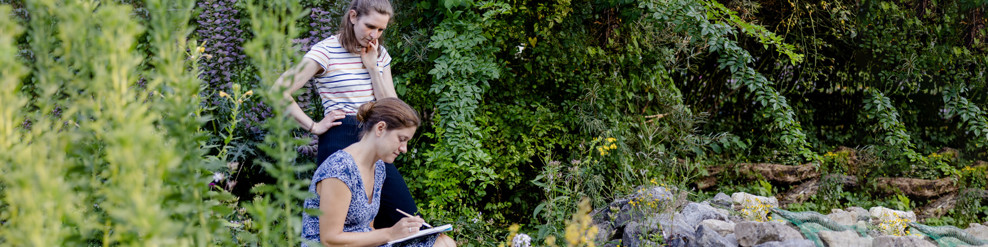 Foto: zwei junge Frauen im Garten des Ökologischen Bildungszentrums bei der Dokumentation von Pflanzen