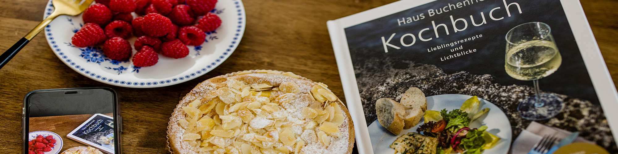 eine Schüssel mit Himbeeren, ein Kuchen, ein Kochbuch und ein Smartphone