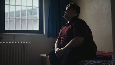 Filmstill aus "Haus 4": deutlich übergewichtige Person sitzend in einer Gefängniszelle