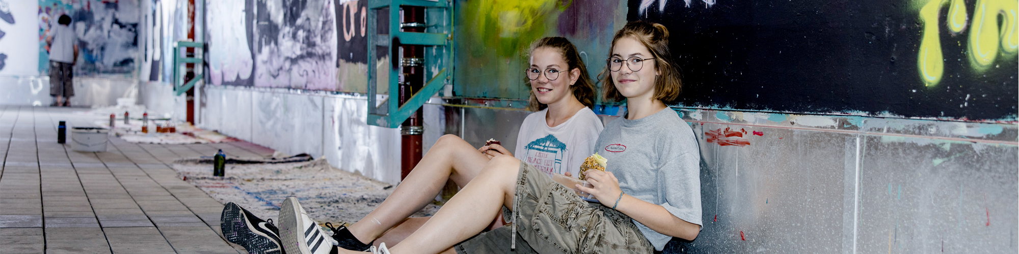 zwei junge Mädchen, sitzend vor einer Wand mit Graffitis
